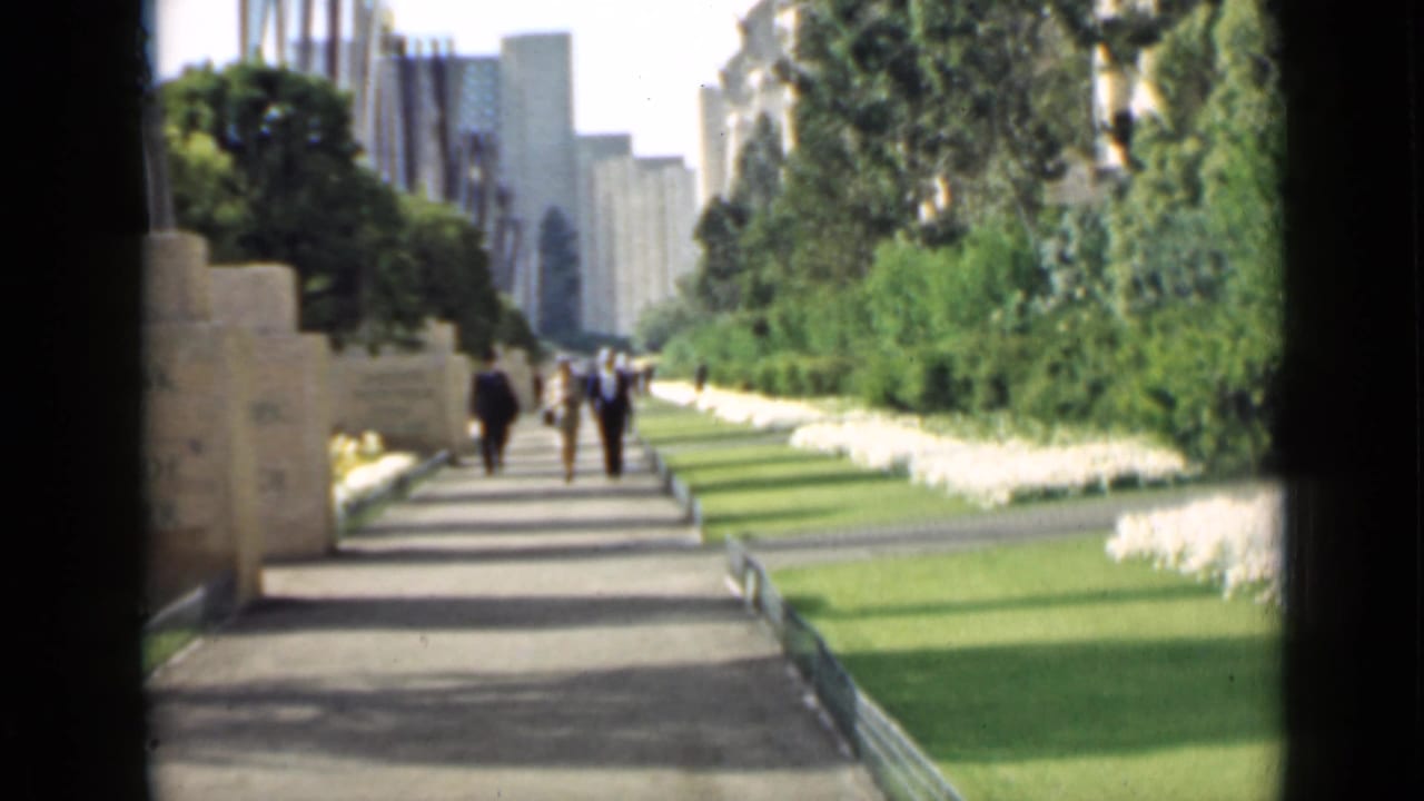 Pedestrians walk along a landscaped path with lawns, white flowers, stone walls, and tall buildings beyond.