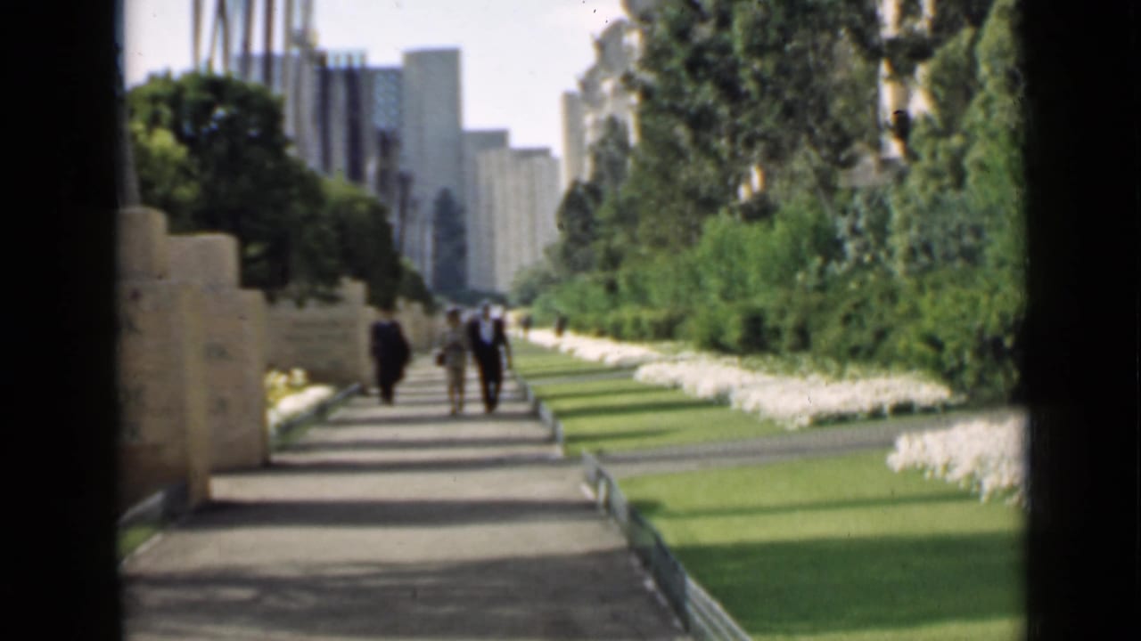Several people walk on a sunlit garden path bordered by lawns, flowers, trees, and stone features.