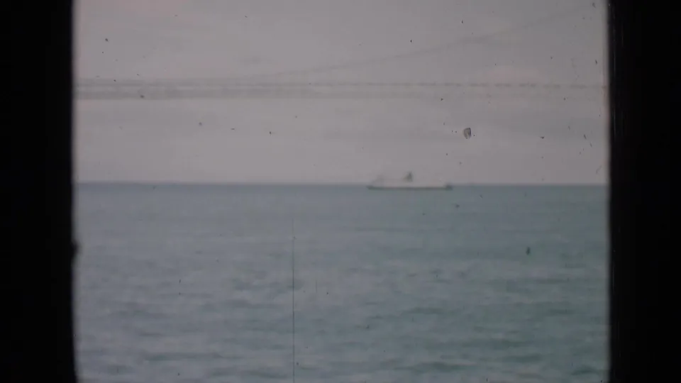 1939 San Francisco home-movie frame showing a small sailboat on calm water, viewed through dark window mullions with hazy sky above.