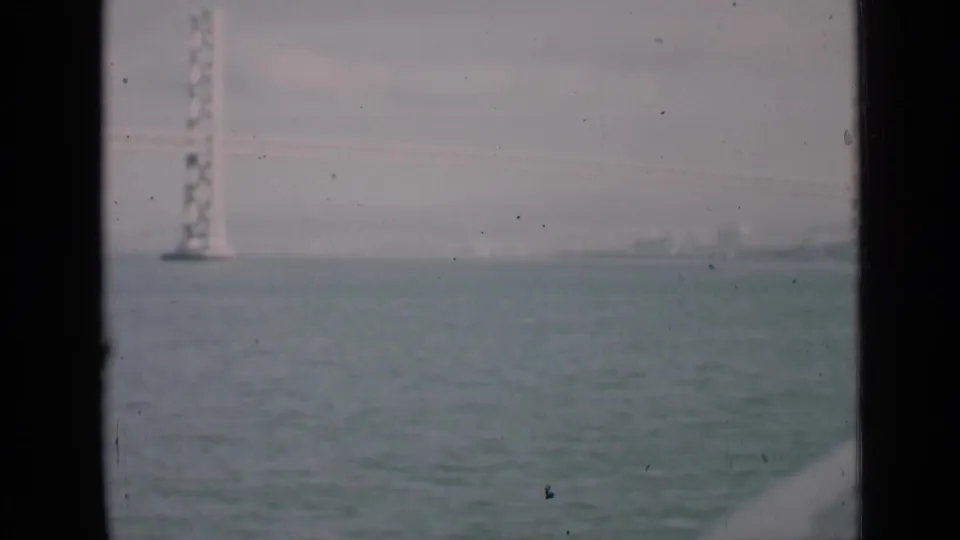 A suspension bridge tower rises over San Francisco water in a 1939 home-movie frame, with distant shoreline structures in haze.