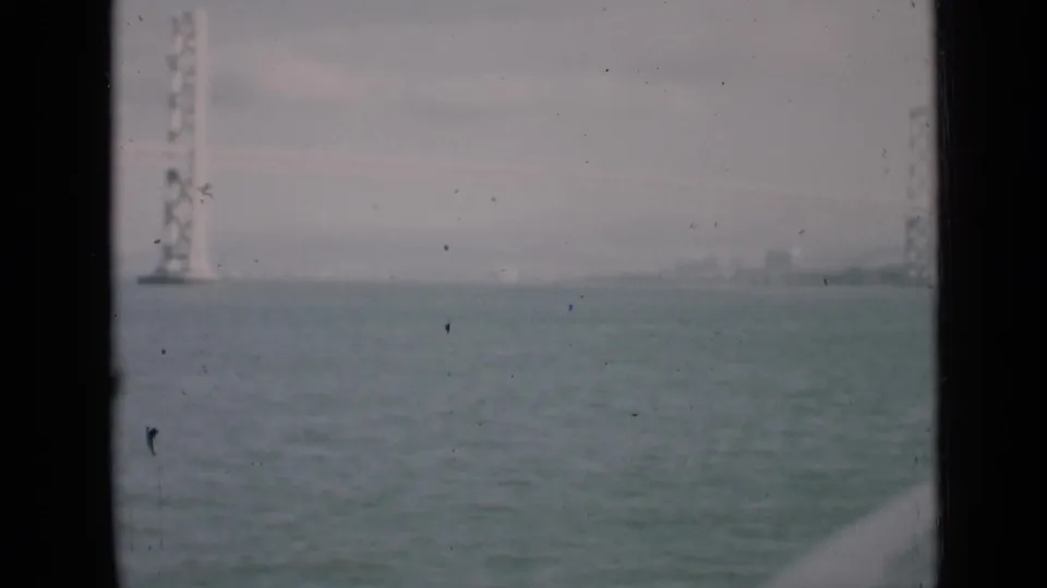 Hazy 1930s waterfront view with a bridge tower at left, distant skyline, and rippled bay surface.