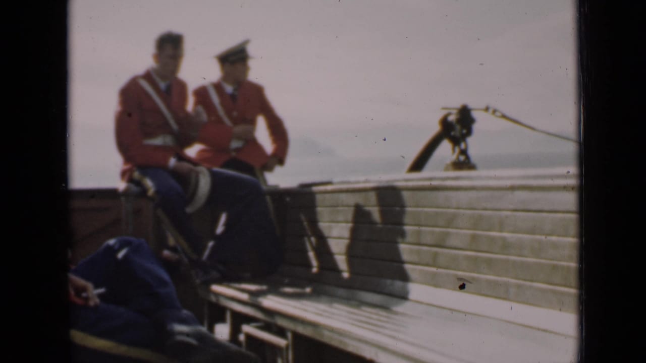 Two uniformed men in red jackets sit on a wooden bench beside a deck rail and rope hardware.
