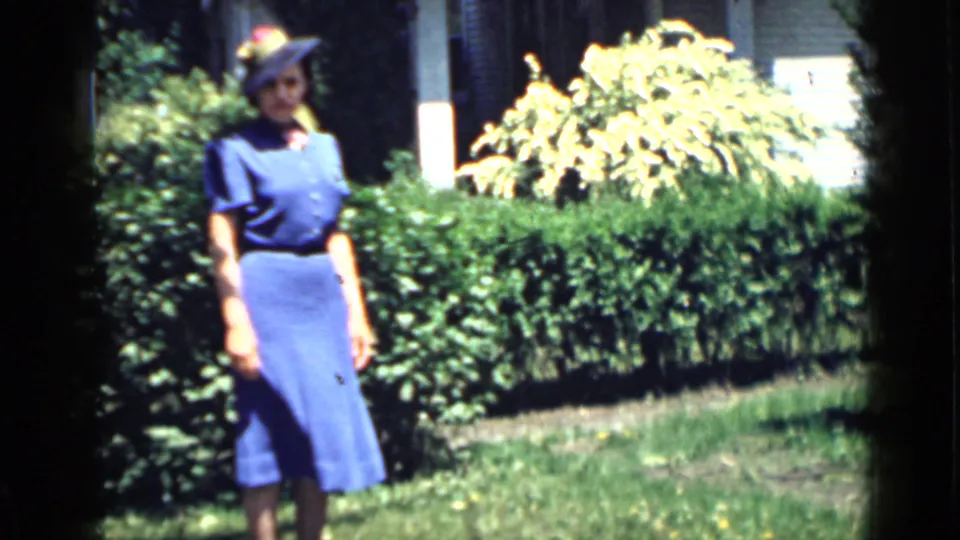 1939 San Francisco home-movie frame of an adult woman in a blue dress and hat standing in a garden before a residential house.