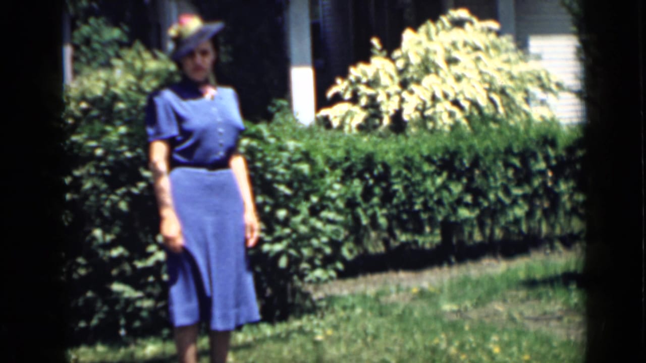 Woman in a blue dress and tilted hat standing in a sunny garden beside a hedge.