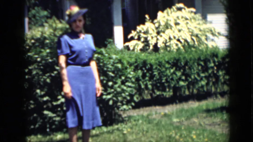 In supported 1939 San Francisco home-movie footage, a woman in a blue dress and hat poses near a hedge and house exterior.