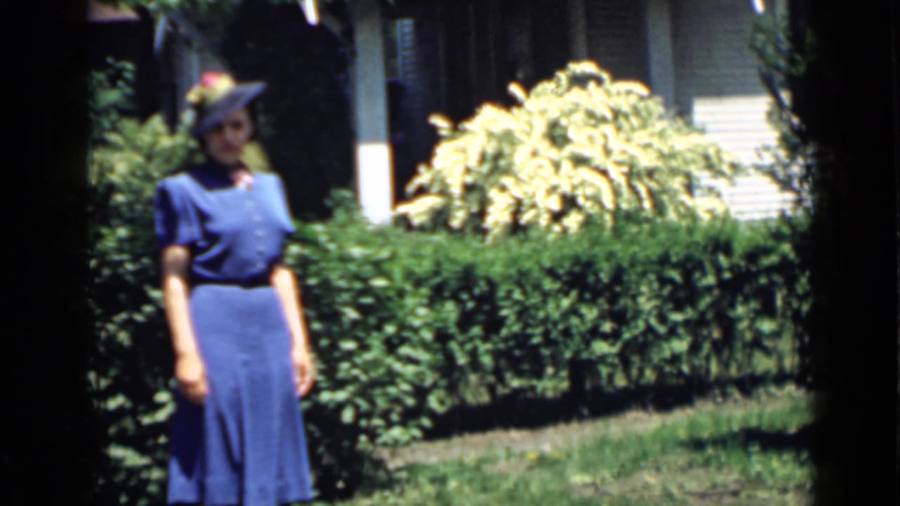 Woman in blue standing before a hedge with sunlit shrubs and a house behind her.