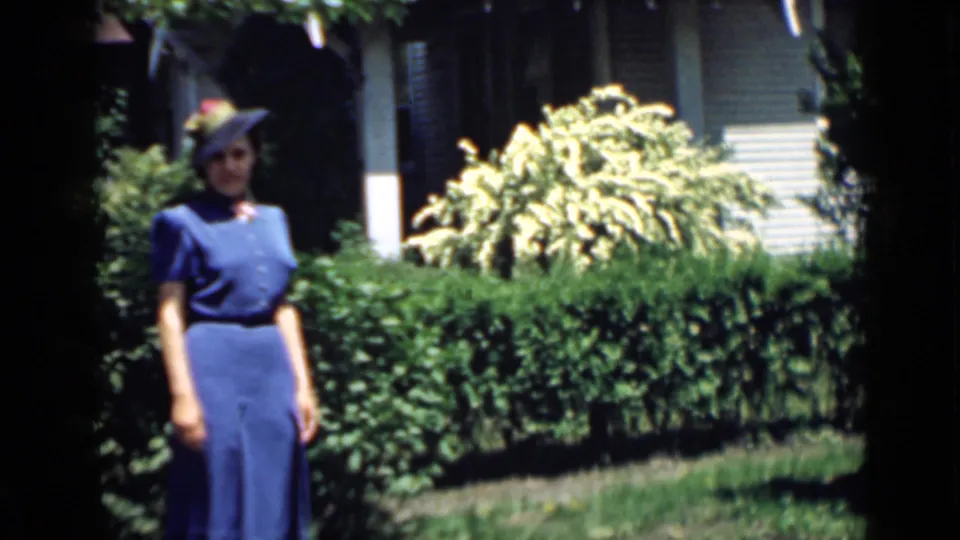 The home-movie frame shows the woman closer to camera, wearing a blue dress and hat while posing in front of a manicured garden.