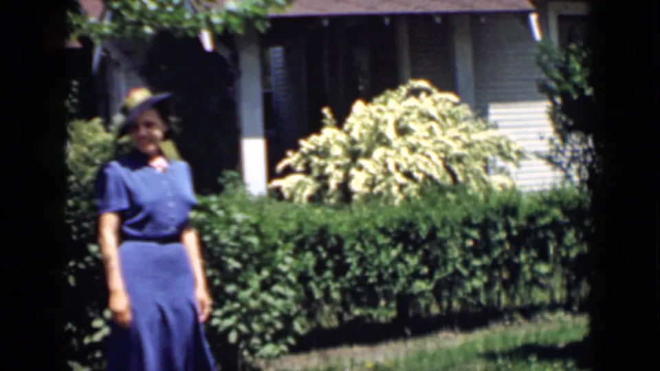 A woman appears to smile while posing in a blue dress and hat in the supported 1939 San Francisco garden scene.