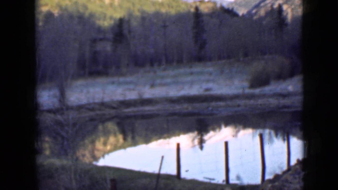 1939 Utah mountain pond view with fence posts near the shore, bare trees, evergreen slopes, and pale reflected sky on the water.