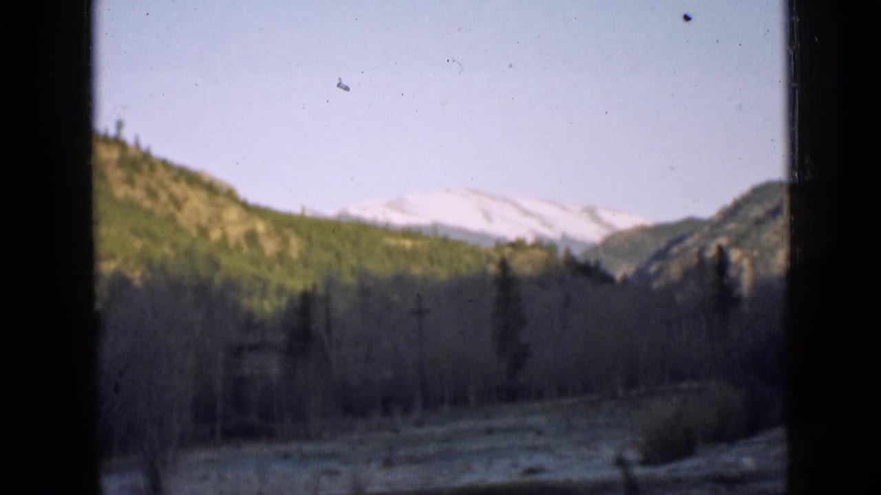 Mountain valley seen through dark window or film-frame edges, with snow-capped peaks beyond forested slopes.