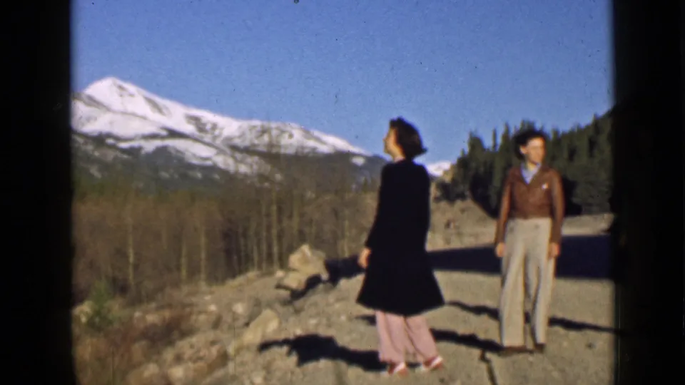 Two women in period clothing stand on a Utah mountain road or overlook in 1939 home-movie footage, backed by snowy peaks and trees.