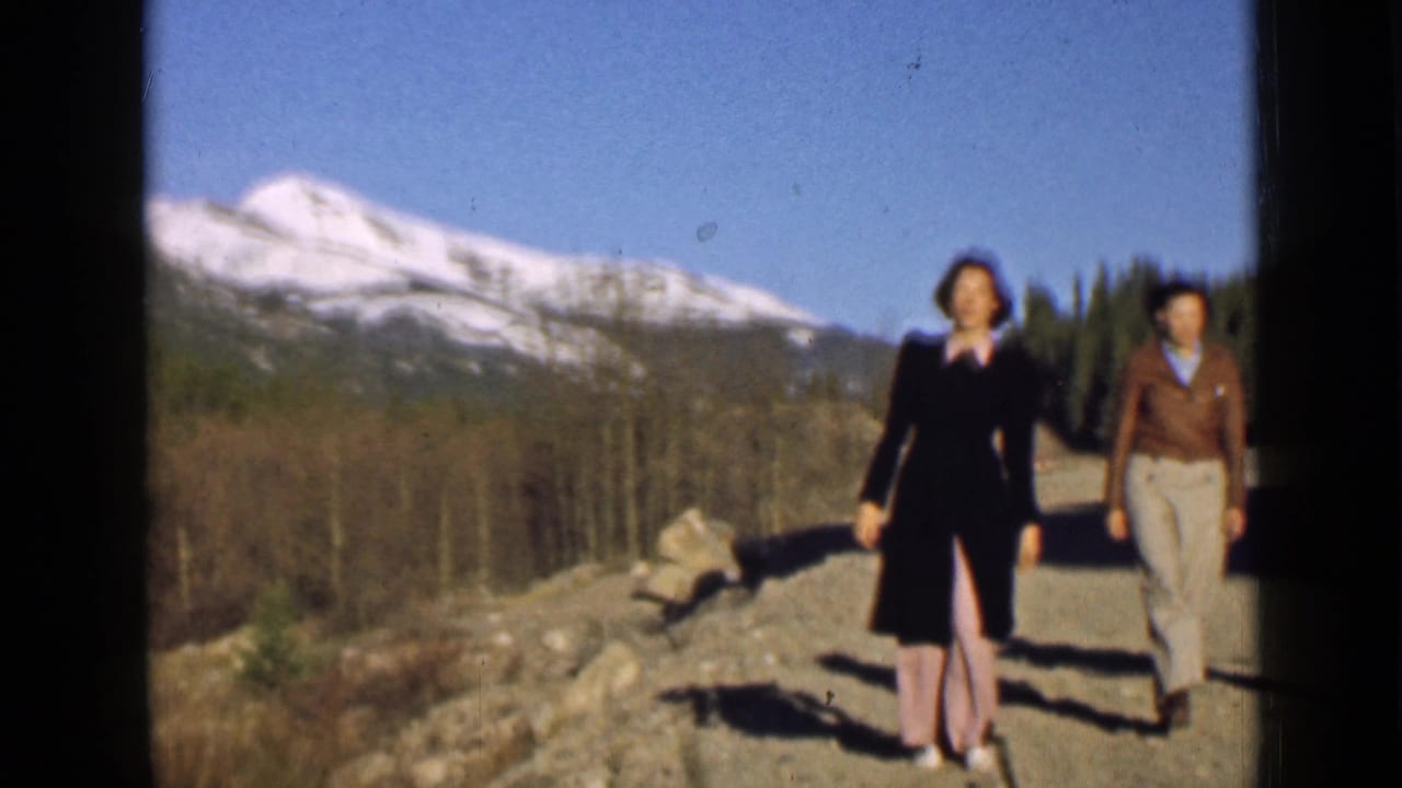 Two women walk on a dirt mountain road with snowy peaks and trees behind them.