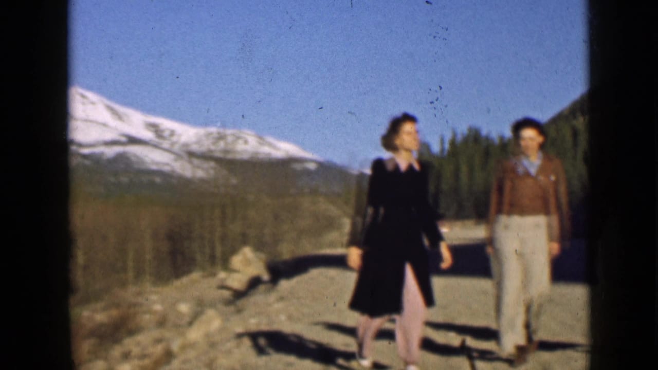 Two women walk side by side on a Utah mountain road, with snowy peaks and dark evergreen forest behind them.