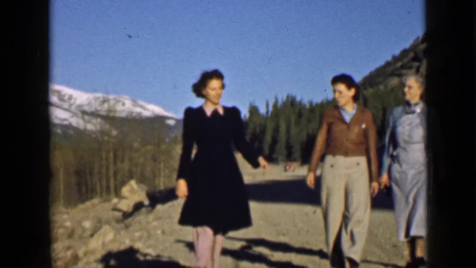 Three women in a 1939 Utah mountain scene walk together on a dirt road beneath snow-capped peaks.