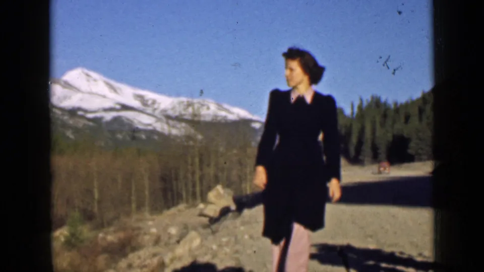 A woman in dark clothing walks past the camera in Utah, with snowy mountains, trees, and road shadows behind her.