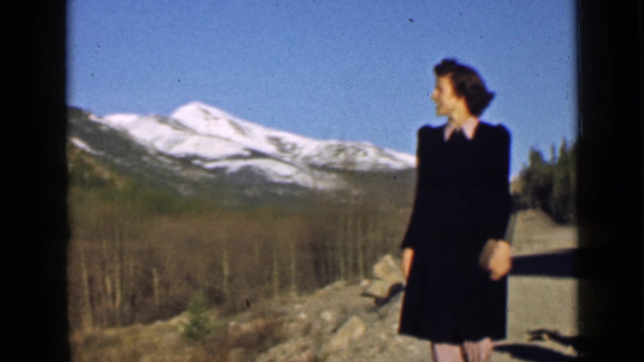 Woman in a dark dress stands near rocks with snow-capped mountains behind her.