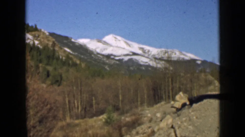 The camera holds on a Utah mountain landscape with snow-capped peaks, bare trees, rocks, and a clear sky.