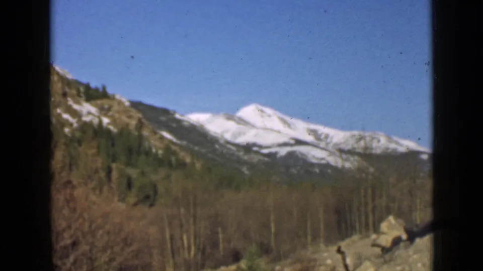 Soft 8mm frame of snow-covered mountain peaks above wooded slopes, with vehicle-window edges visible in the foreground.