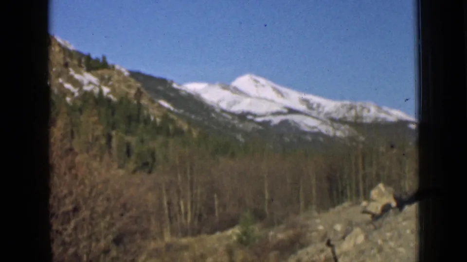 Archival 8mm view of snow-capped peaks, forested slopes, bare trees, and roadside rocks from a traveler’s window perspective.
