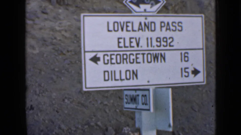 Home-movie frame of the Loveland Pass roadside sign, with Summit County marker below and rocky slope behind it.