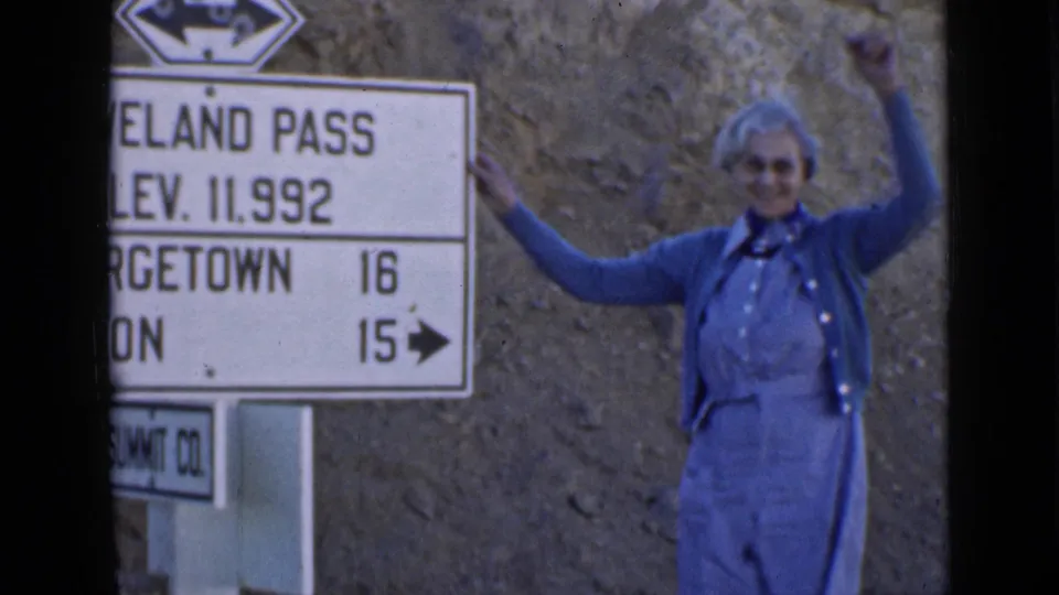 An elderly woman in blue clothing raises her arms beside the Loveland Pass elevation sign in 1930s home-movie footage.