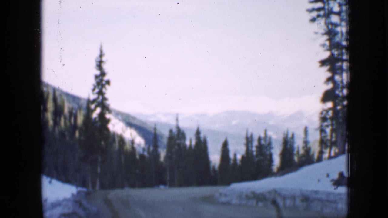 1939 Utah home-movie frame of a snow-lined forest road curving between tall conifers, with hazy mountain ridges beyond.