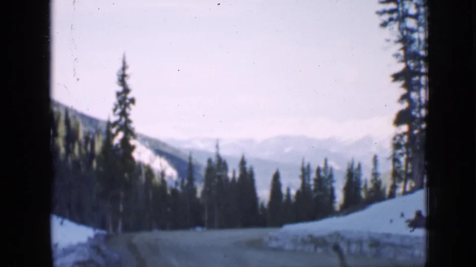 1939 Utah home-movie frame of a snow-lined forest road curving between tall conifers, with hazy mountain ridges beyond.