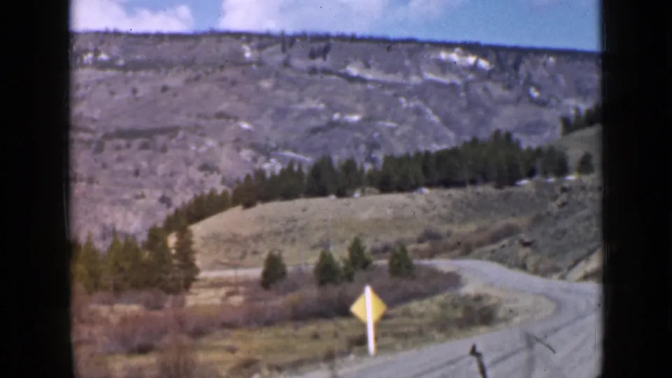 1939 Utah 8mm frame showing a winding road leaving forest for an open hillside with a small yellow road sign.