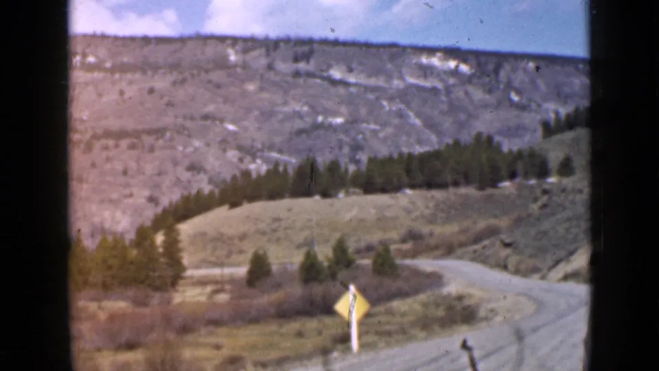 1939 Utah home-movie frame showing a dirt road curving downhill past a yellow warning sign, with evergreens and rocky hillsides beyond.