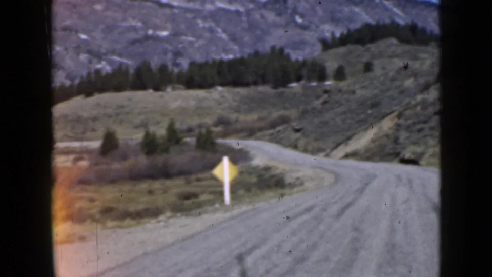1939 Utah home-movie view of a downhill dirt road bend with a yellow warning sign, open grassland, evergreens, and rocky slopes.