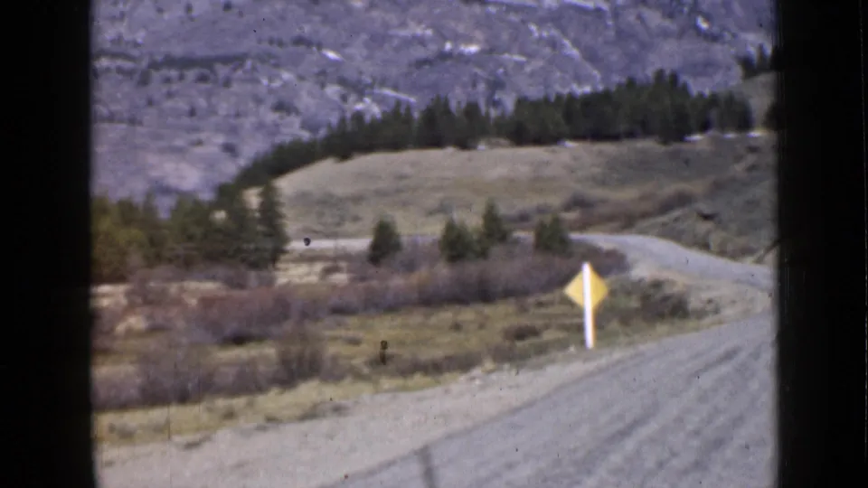 1939 Utah frame showing the winding dirt road from a vehicle perspective, with a yellow sign, scattered trees, and rugged mountain backdrop.