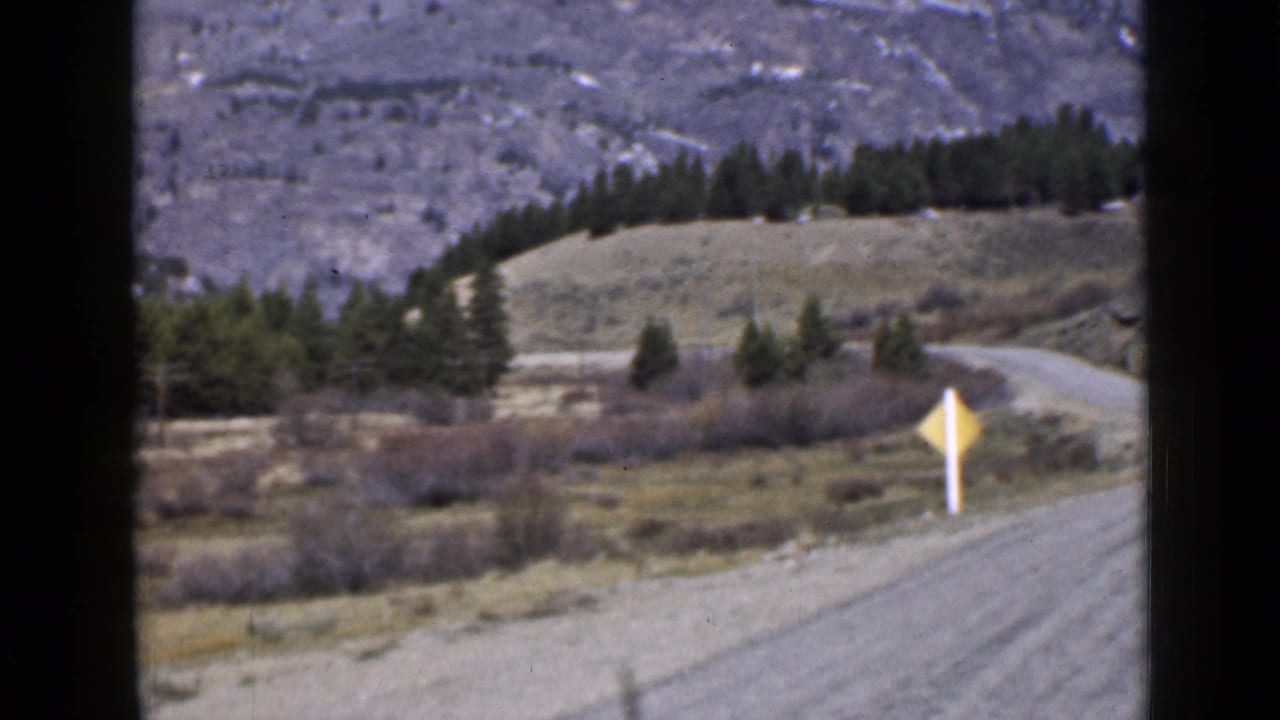 Winding dirt road with a yellow warning sign, trees, hills, and distant grey mountains.