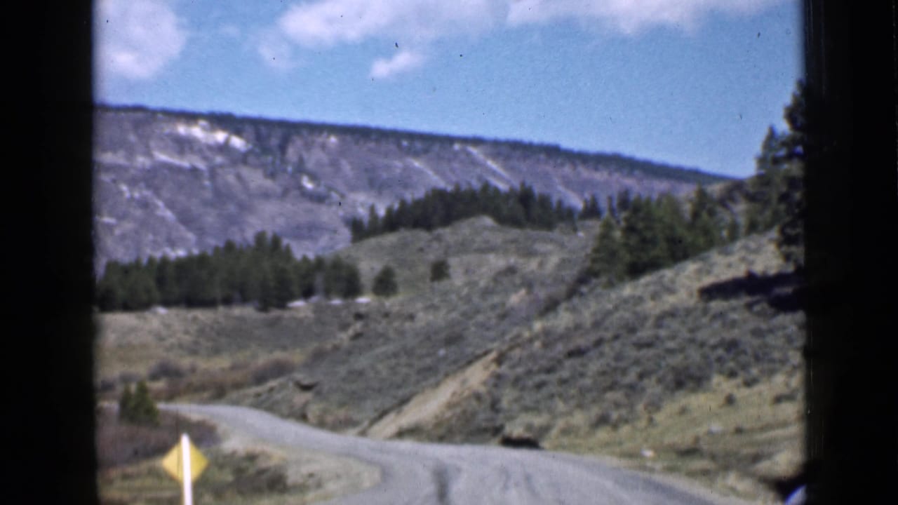 Road curving through dry hills with trees and black film borders.