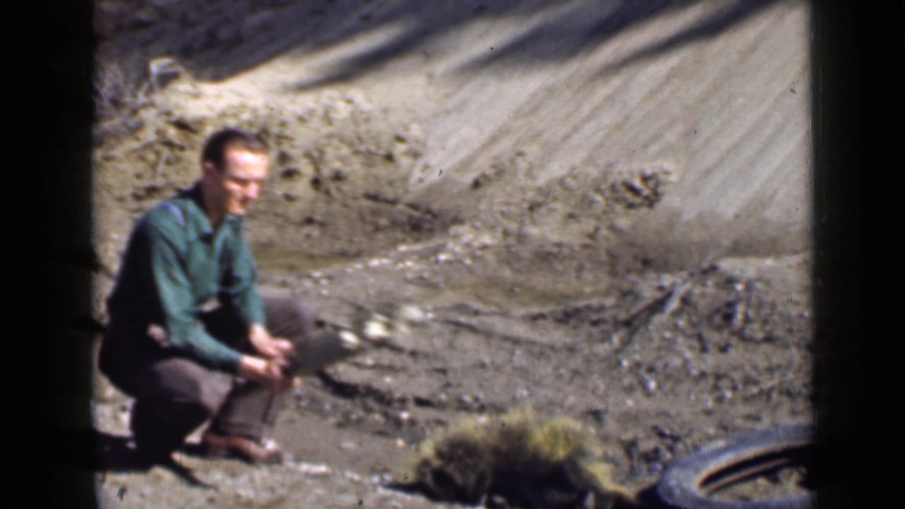 1939 Utah 8mm frame showing an adult man kneeling beside a wooden plank in a dry roadside or hillside setting.