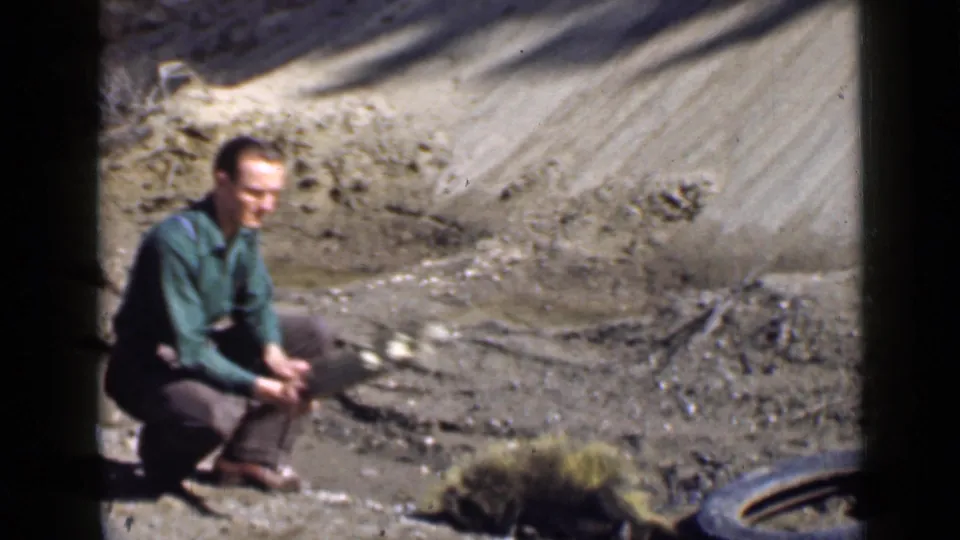 1939 Utah 8mm frame showing an adult man kneeling beside a wooden plank in a dry roadside or hillside setting.