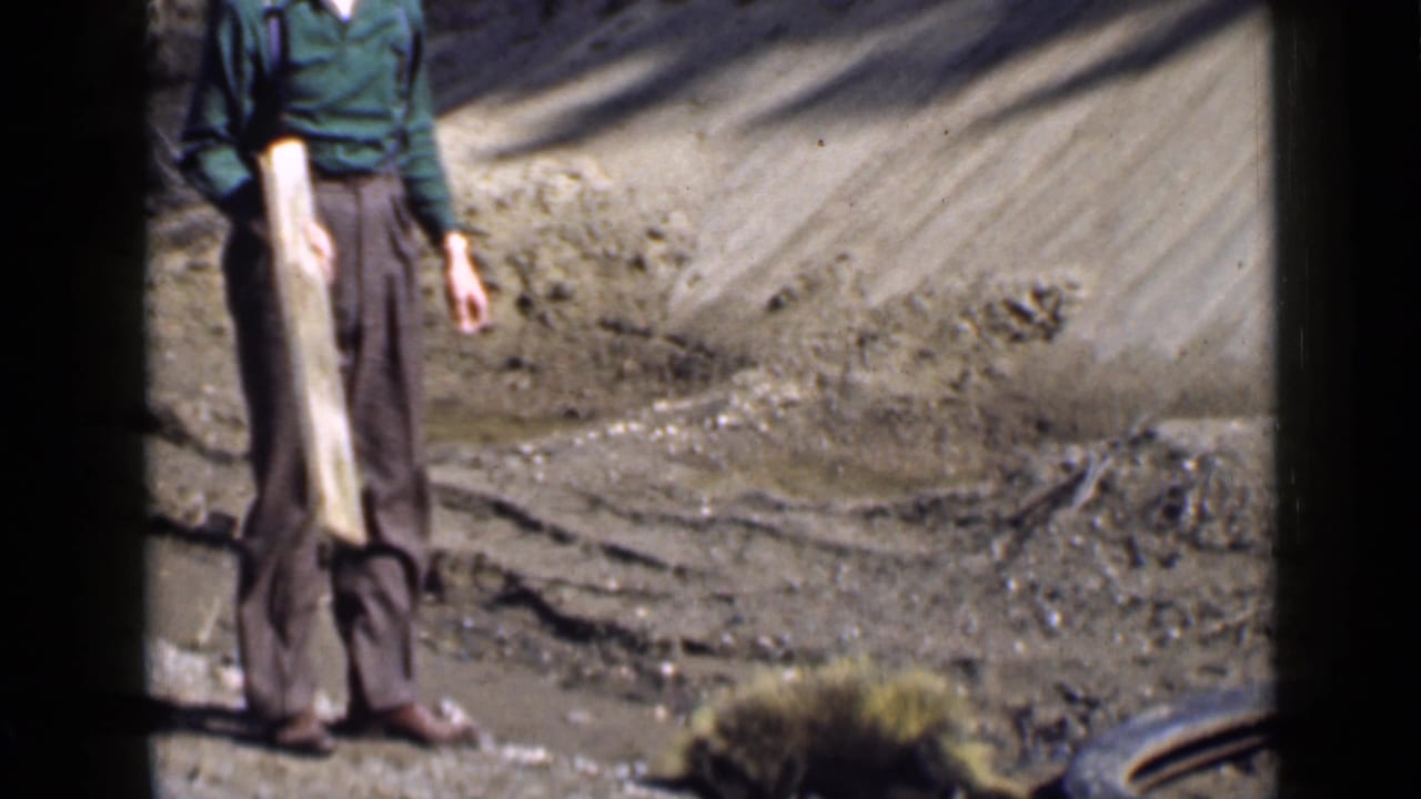 1939 Utah home-movie frame of an adult man standing with a long wooden plank near a dry embankment and sparse vegetation.