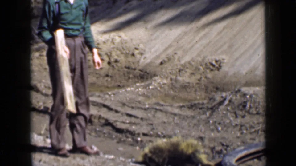 1939 Utah home-movie frame of an adult man standing with a long wooden plank near a dry embankment and sparse vegetation.