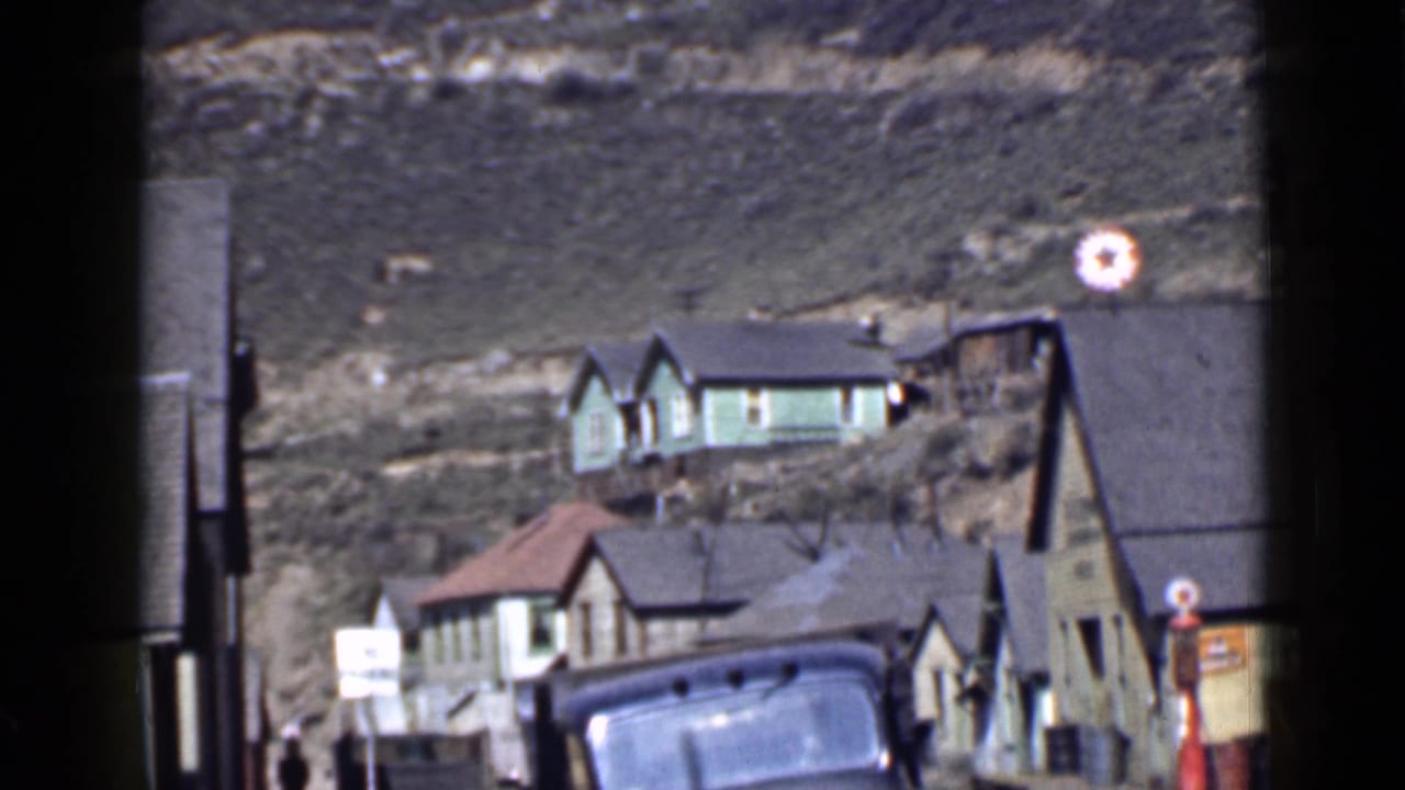 1939 Utah exterior view with a green hillside house, neighboring rooftops, signs, and a partial vehicle in a sunny small-town scene.