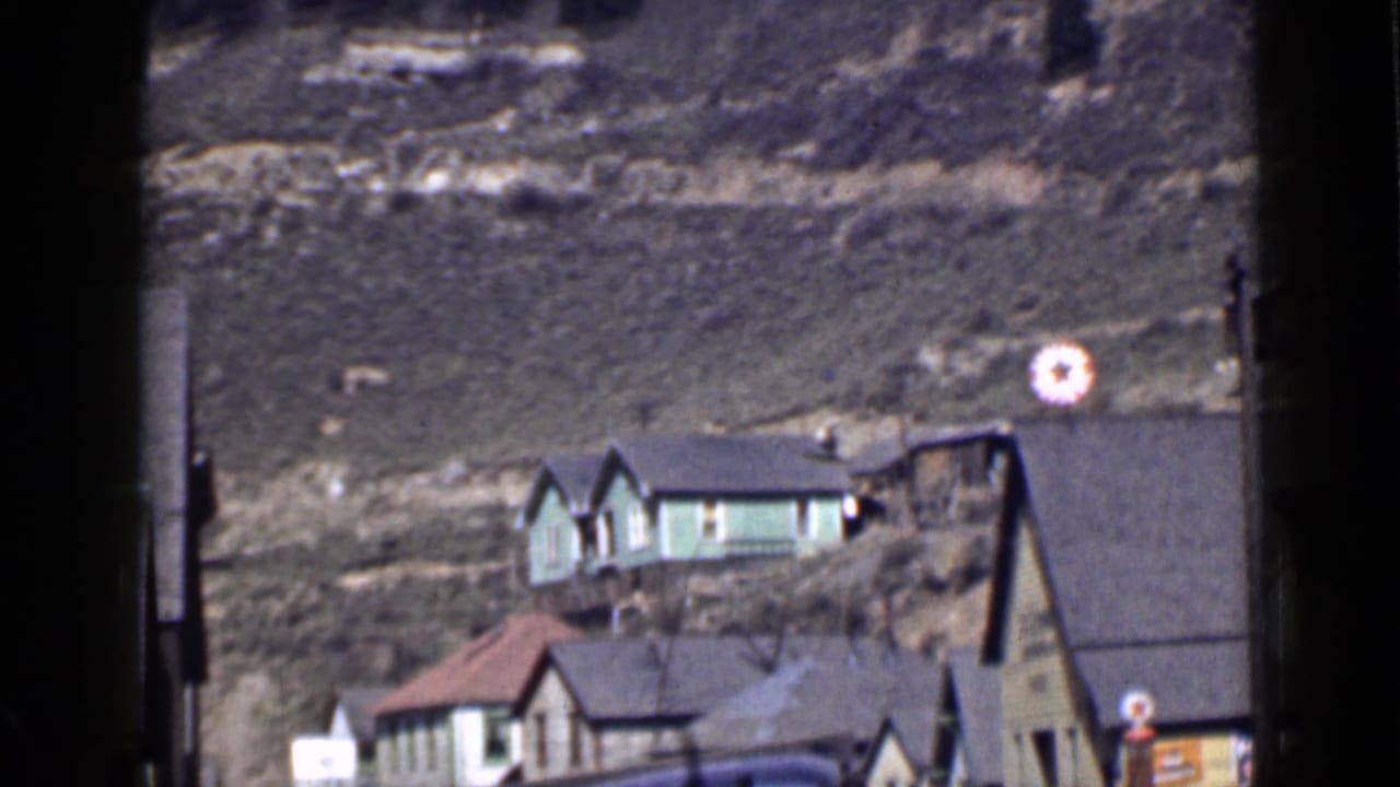 1939 Utah home-movie view of small houses below a grassy hill, with a green house higher on the slope and roadside details below.