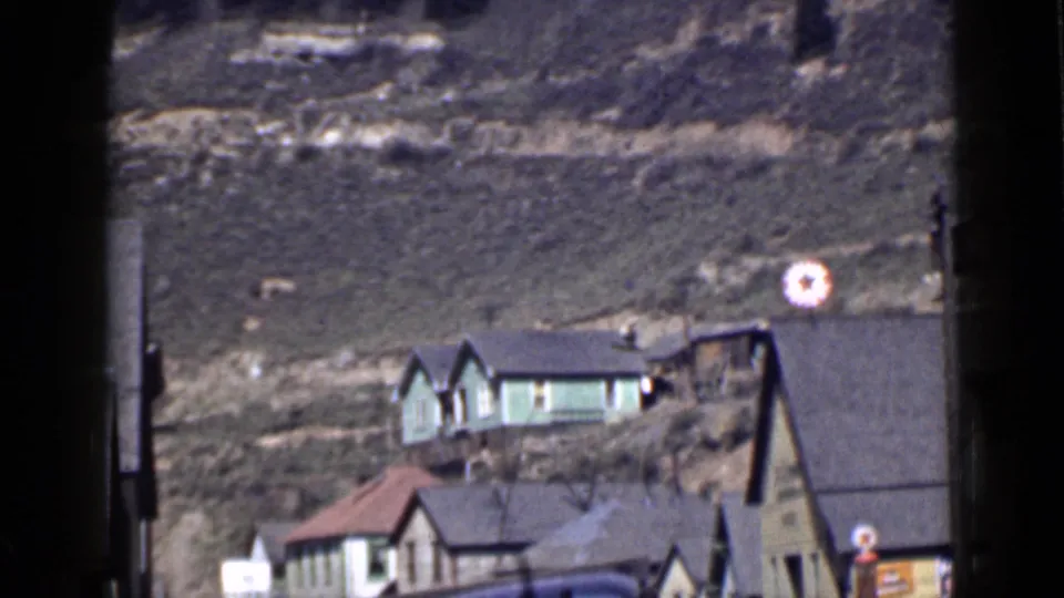 1939 Utah home-movie view of small houses below a grassy hill, with a green house higher on the slope and roadside details below.