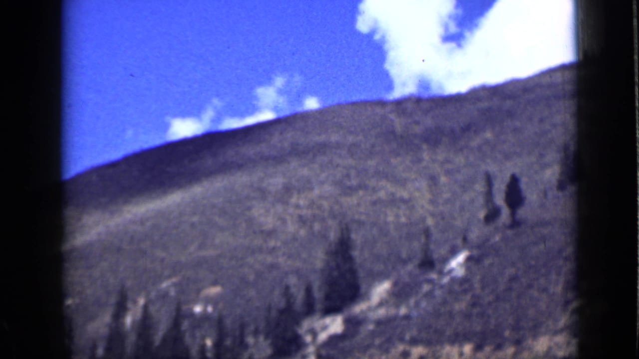 1939 Utah 8mm frame of a sunlit hillside with scattered evergreens, blue sky, and a pale snow or rock patch.
