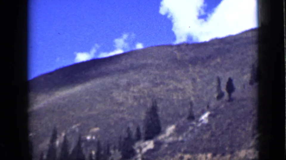 1939 Utah 8mm frame of a sunlit hillside with scattered evergreens, blue sky, and a pale snow or rock patch.