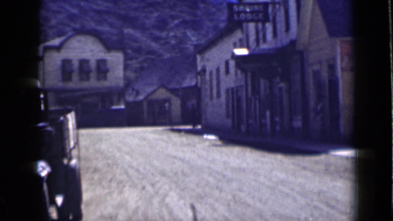 Quiet dirt street with storefront buildings, parked automobiles, and a Shrine Lodge sign.