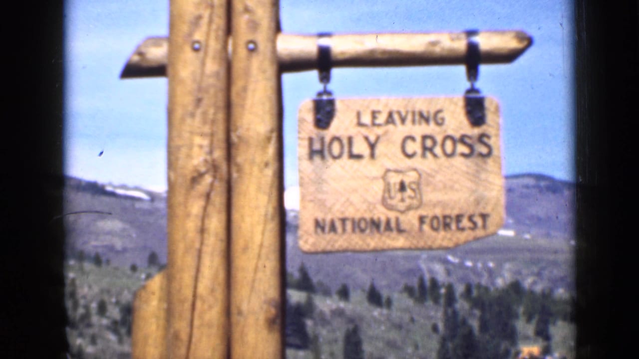1939 Utah frame of a wooden Leaving Holy Cross National Forest sign with U.S. Forest Service shield, rolling hills, sparse trees, and blue sky.