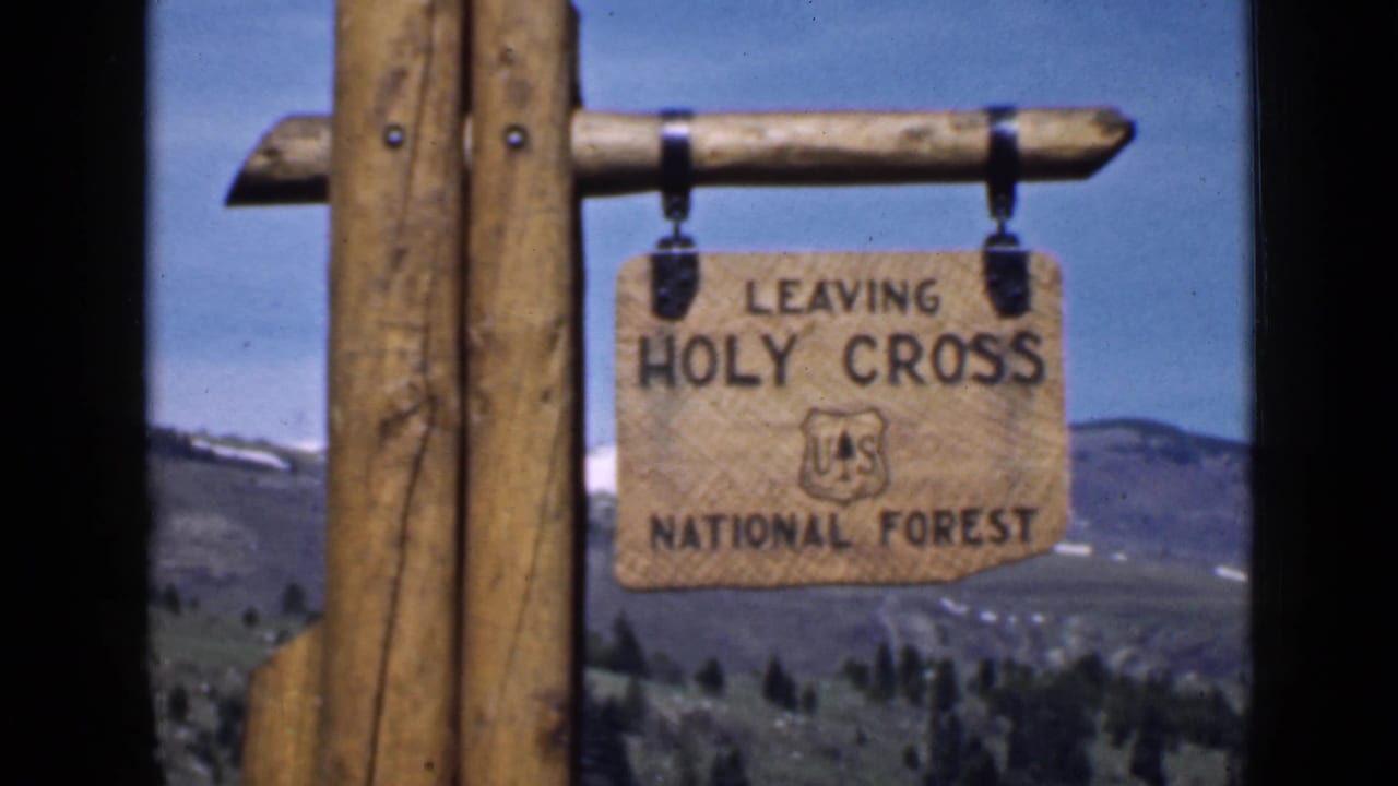 A traveler’s 1939 Utah view of the Leaving Holy Cross National Forest sign suspended from wood posts against hills and open sky.