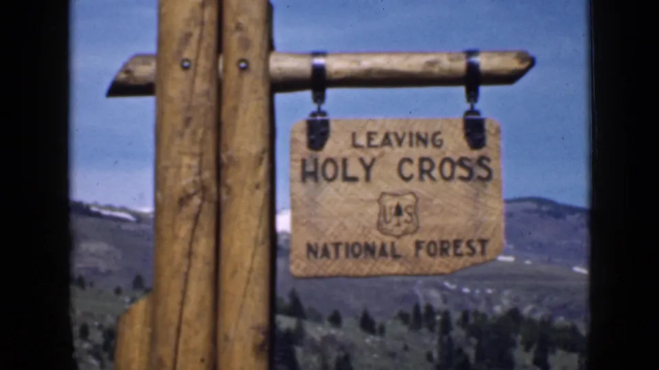 A traveler’s 1939 Utah view of the Leaving Holy Cross National Forest sign suspended from wood posts against hills and open sky.