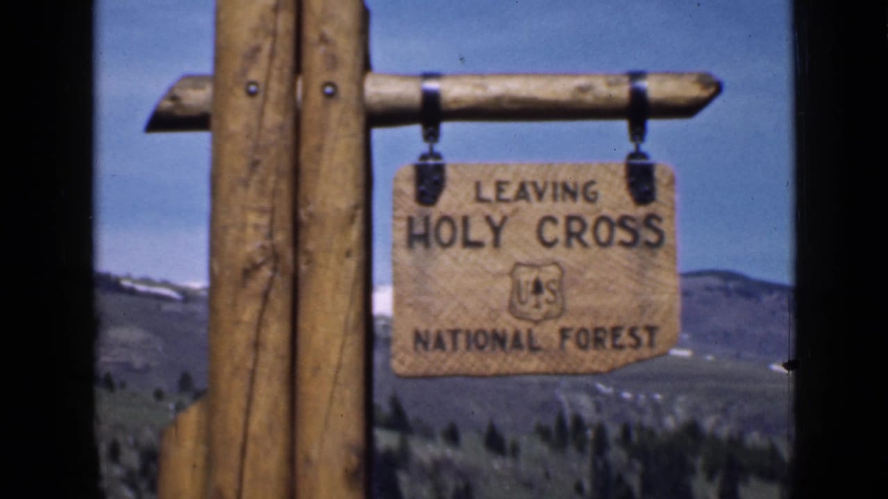 The camera holds on the wooden Holy Cross National Forest exit sign, with the agency emblem and mountain landscape visible behind it.