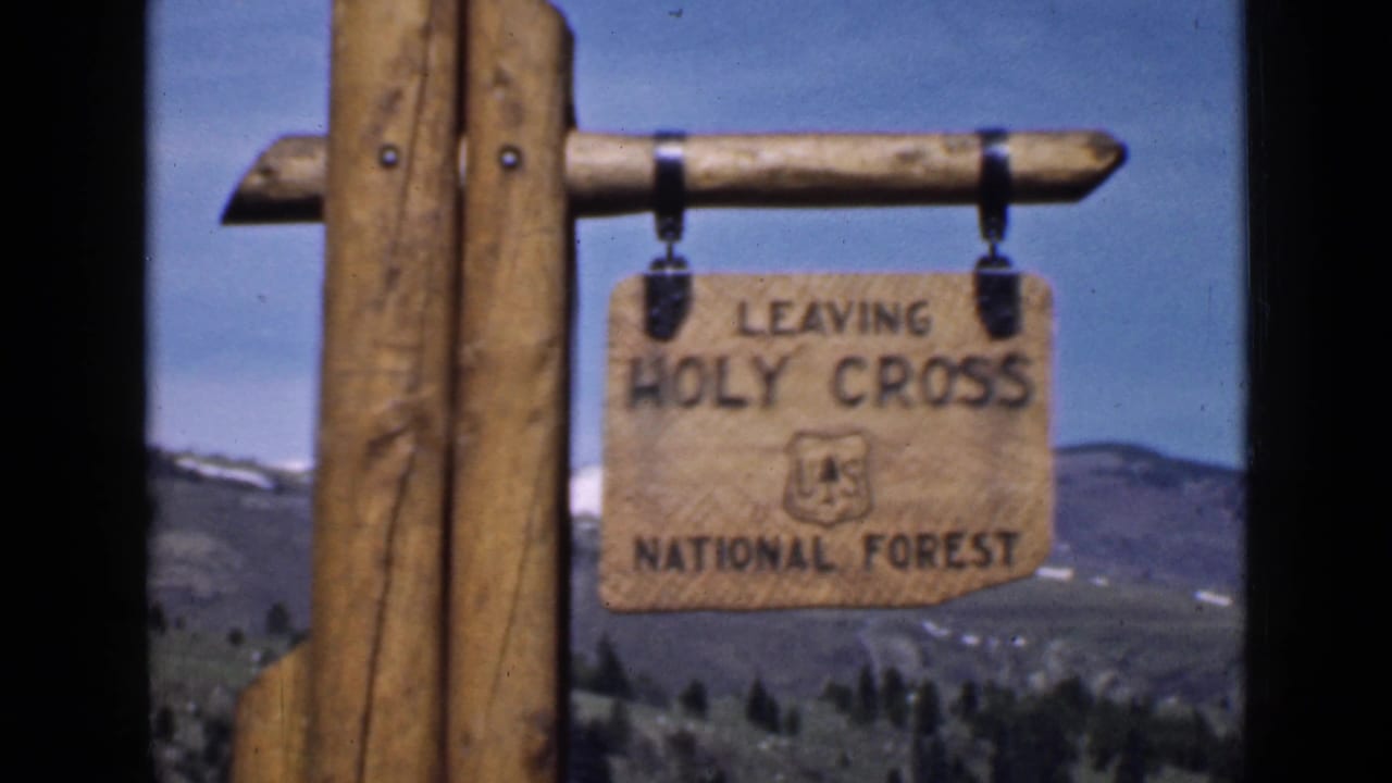 A closer handheld frame centers the wooden forest boundary sign, with the U.S. Forest Service emblem and distant hills still visible.