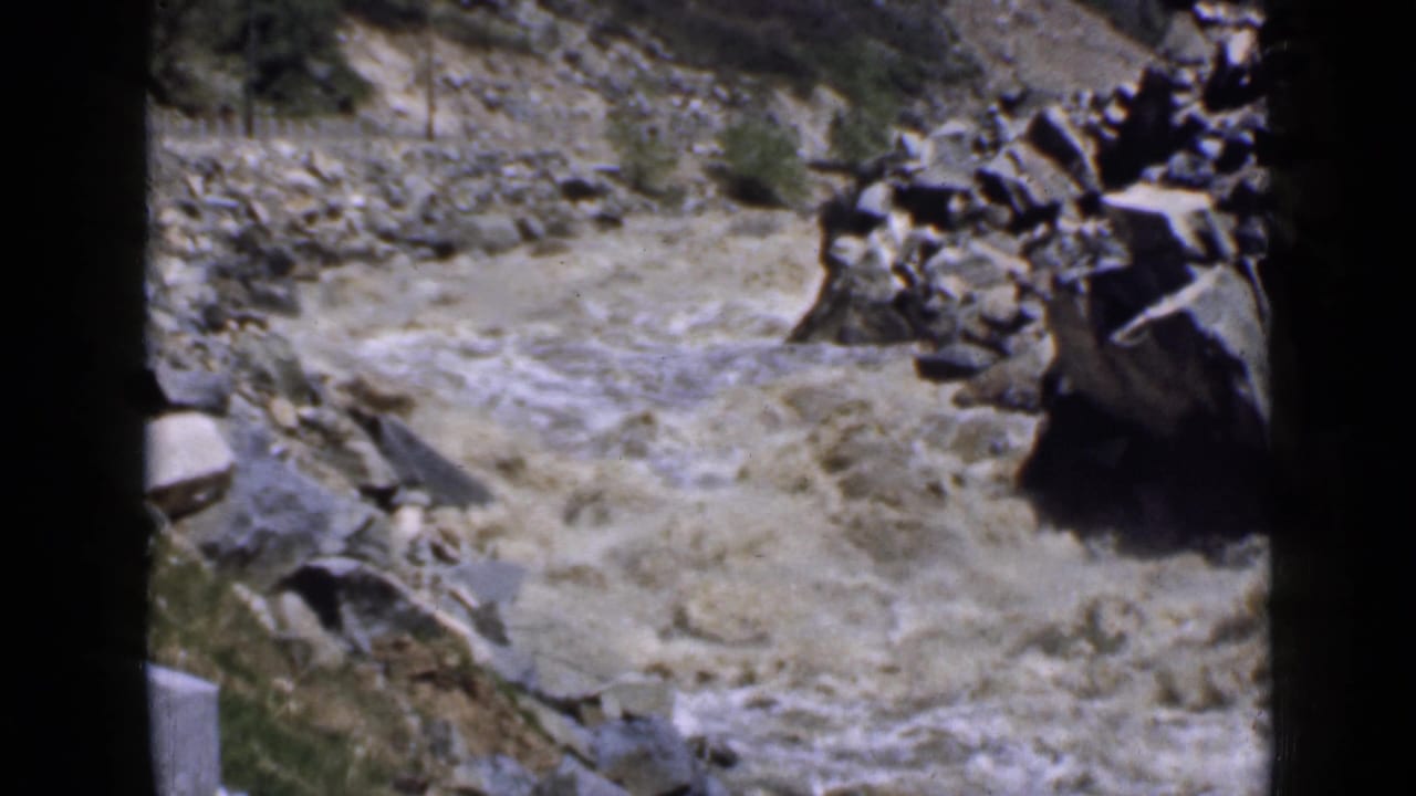Fast muddy water flows between rocky banks and boulders in a canyon.