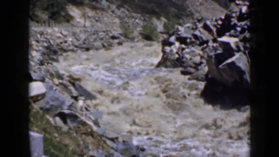 Utah 1939 home-movie frame of a muddy river coursing through rocks, with dry slope and sparse vegetation behind it.